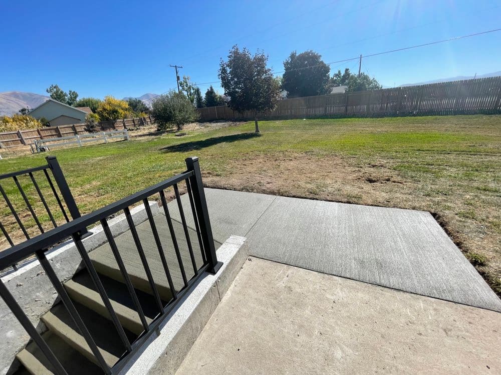 View of a grassy yard with trees and a concrete patio steps under a clear blue sky.