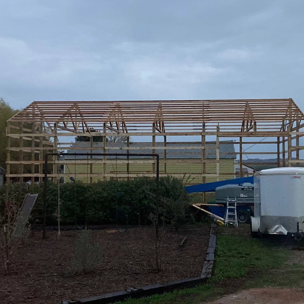 Construction of a wooden barn frame under a cloudy sky, with equipment visible nearby.