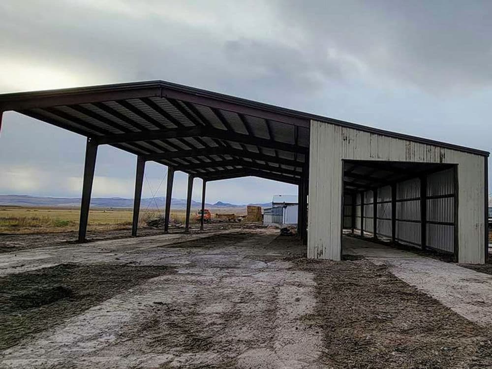 Open metal barn structure on a dirt lot with mountains in the background under cloudy skies.