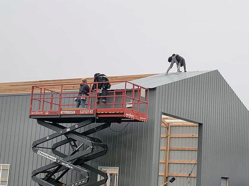 Workers installing metal roofing on a grey building using a scissor lift.