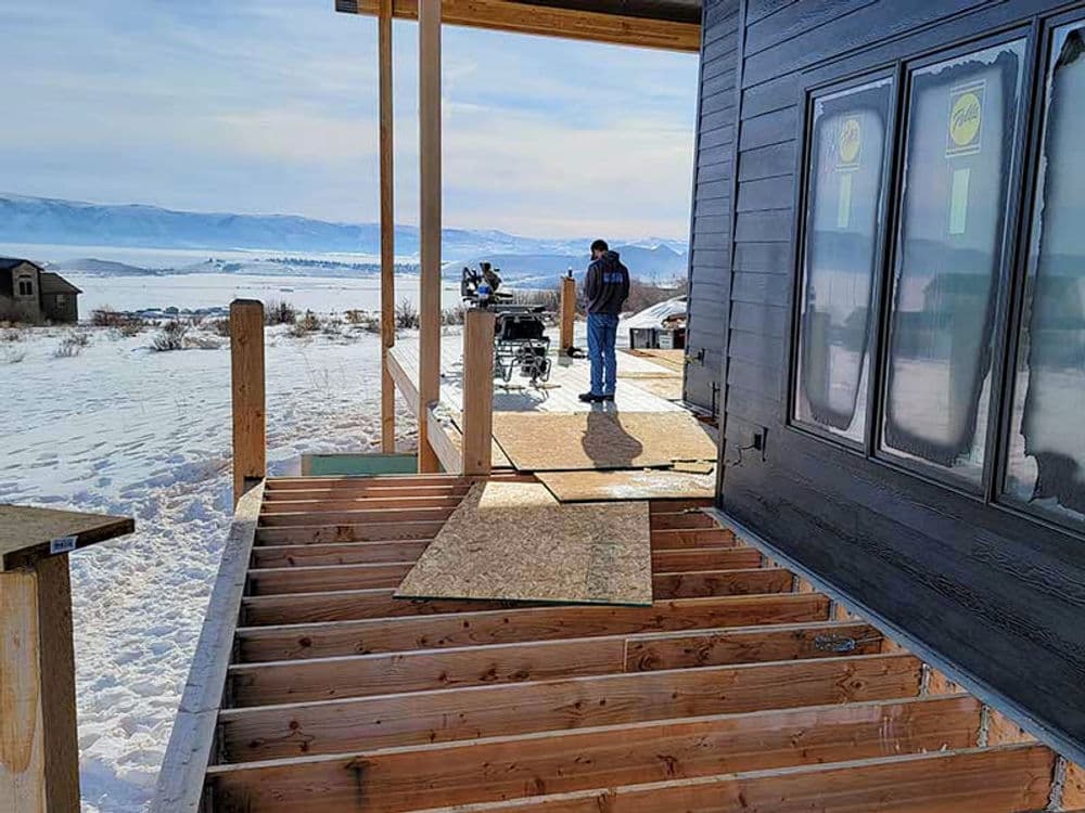 Construction worker on snowy site upgrading deck of a mountain home with scenic views.