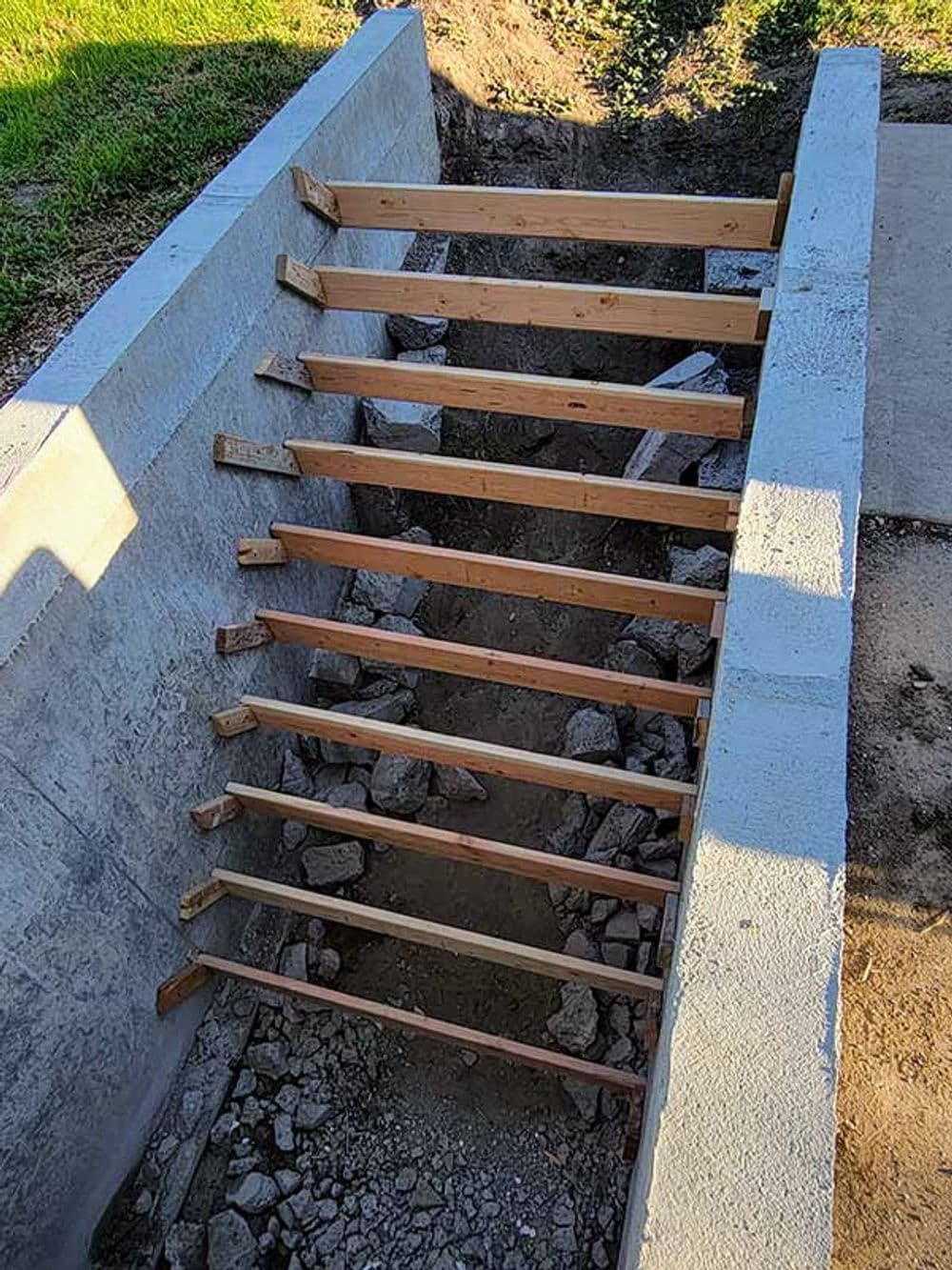 Concrete staircase with wooden steps leading down into a dug-out area, surrounded by grass.